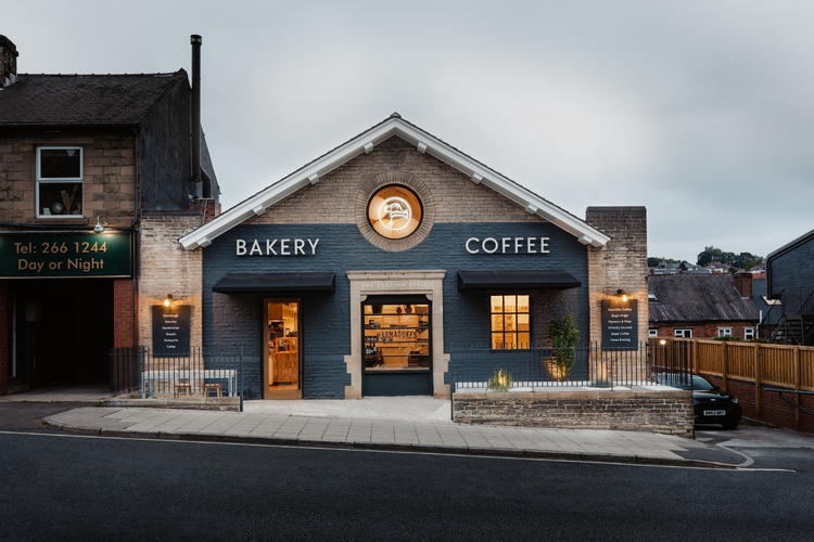  Markadukes shop front, building with pitched roof, warm glow coming from inside. BAKERY COFFEE signs on the front.